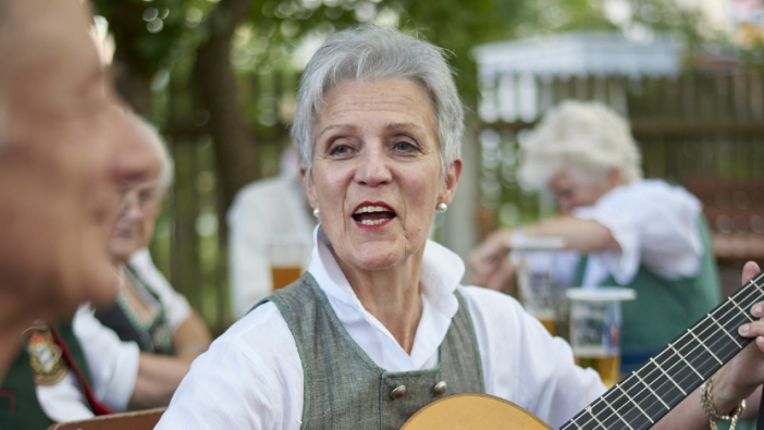 - Eine ältere Frau sitzt im Freien, spielt auf ihrer Gitarre und singt dazu.