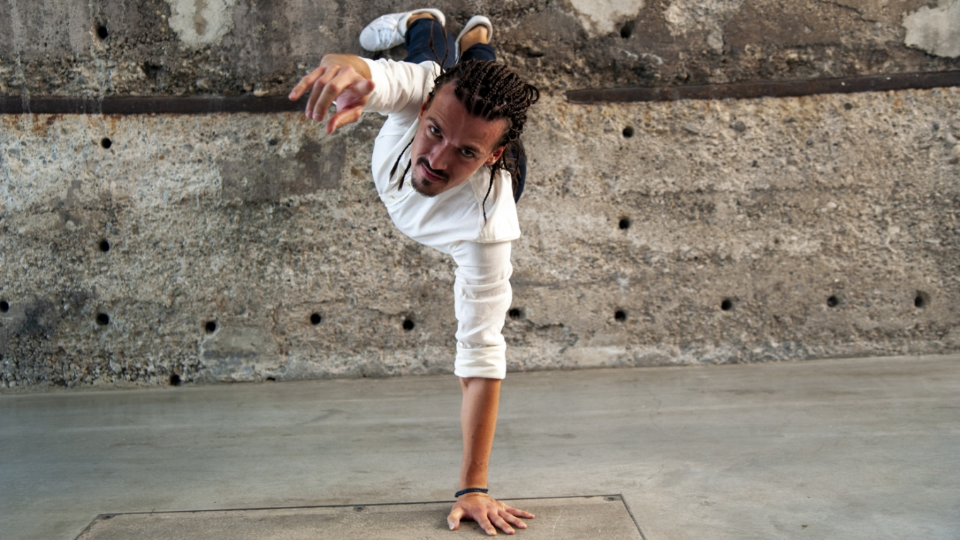 Ein junger Tänzer steht mit den Füßen an die Wand mit nur einer Hand am Boden und zeigt mit der zweiten Hand zur Kamera.