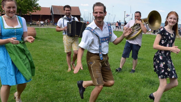 Auf einer Wiese tanzen ein Mann und zwei junge Frauen, im Hintergrund spielt ein Mann Ziach und ein anderer Helikon.