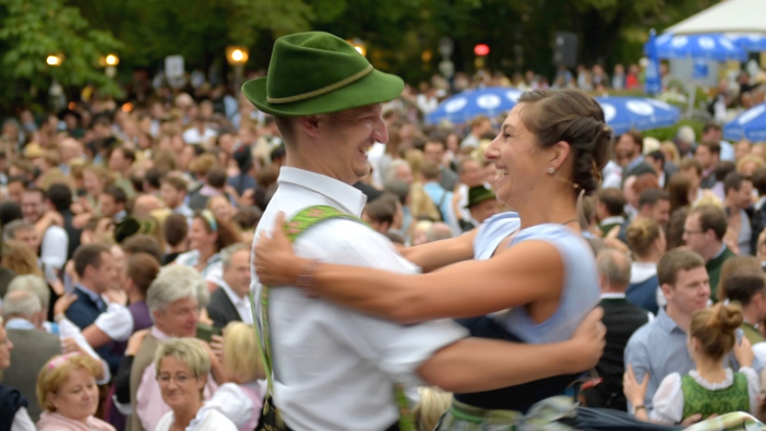 Im Vordergrund tanzen die Tanzmeisterin Katharina Mayer und der Tanzmeister Magnus Kaindl auf einer Bühne miteinander, beide tragen Tracht. Im Hintergrund tanzt eine große Menge Menschen mit.