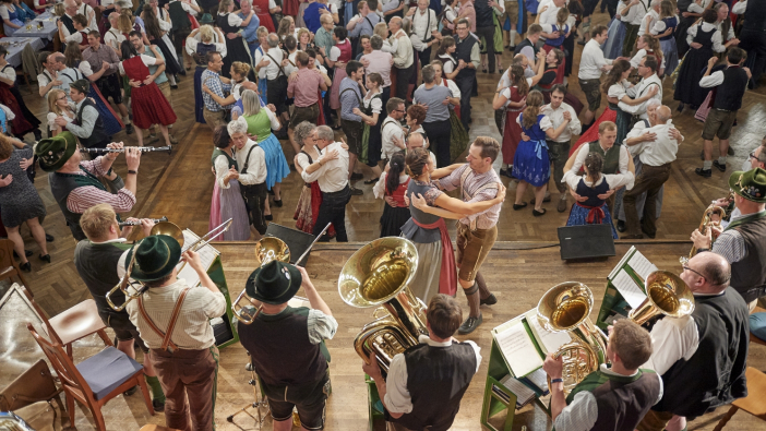 - Zahlreiche lachende Tanzpaare in Freizeitkleidung unterschiedlichen Alters tanzen in einem Saal zu Blasmusik.