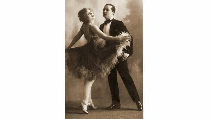 Ein altes schwarzweiß Foto von einem Tanzpaar, sie trägt ein Ballkleid, er einen schwarzen Anzug. Die Frau blickt lächelnd zur Kamera.