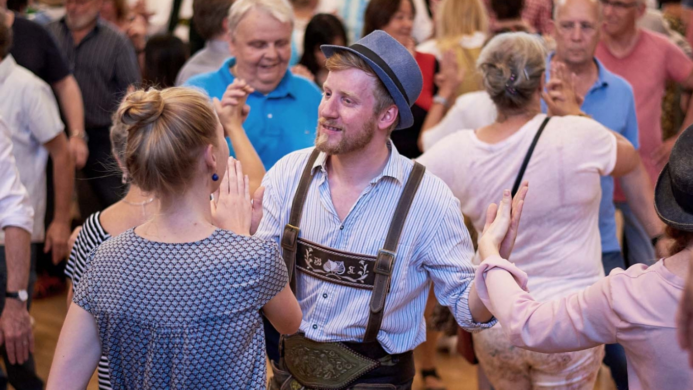 Im schönen Saal des Rathauses tanzen viele Menschen miteinander, in der Mitte ein junges Paar, sie im blauen Kleid und er in Lederhosn.