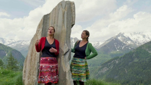 An einem Felsen stehen eine Frau in rot und eine in grün gekleidet und singen miteinander.