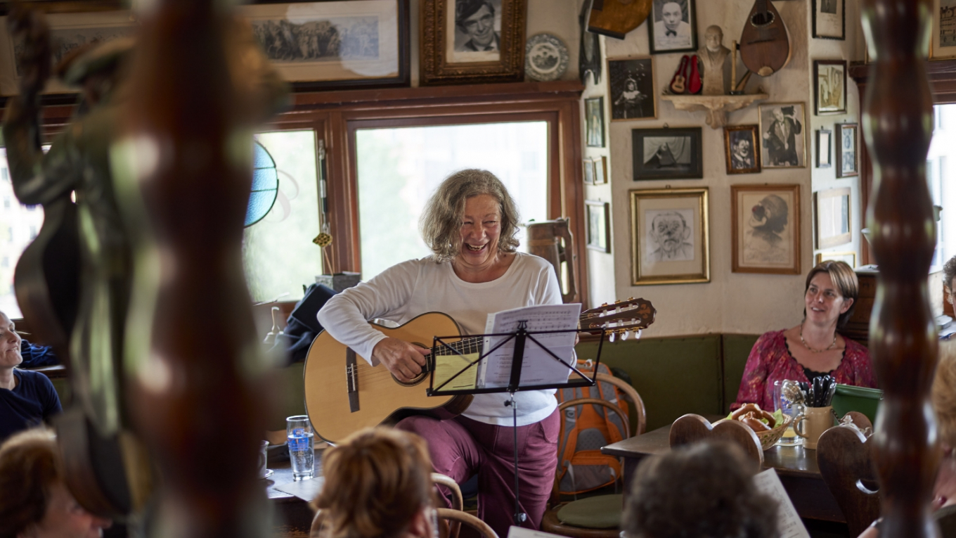 Eine Frau mit Gitarre sitzt umringt von Menschen in einem Café und singt.