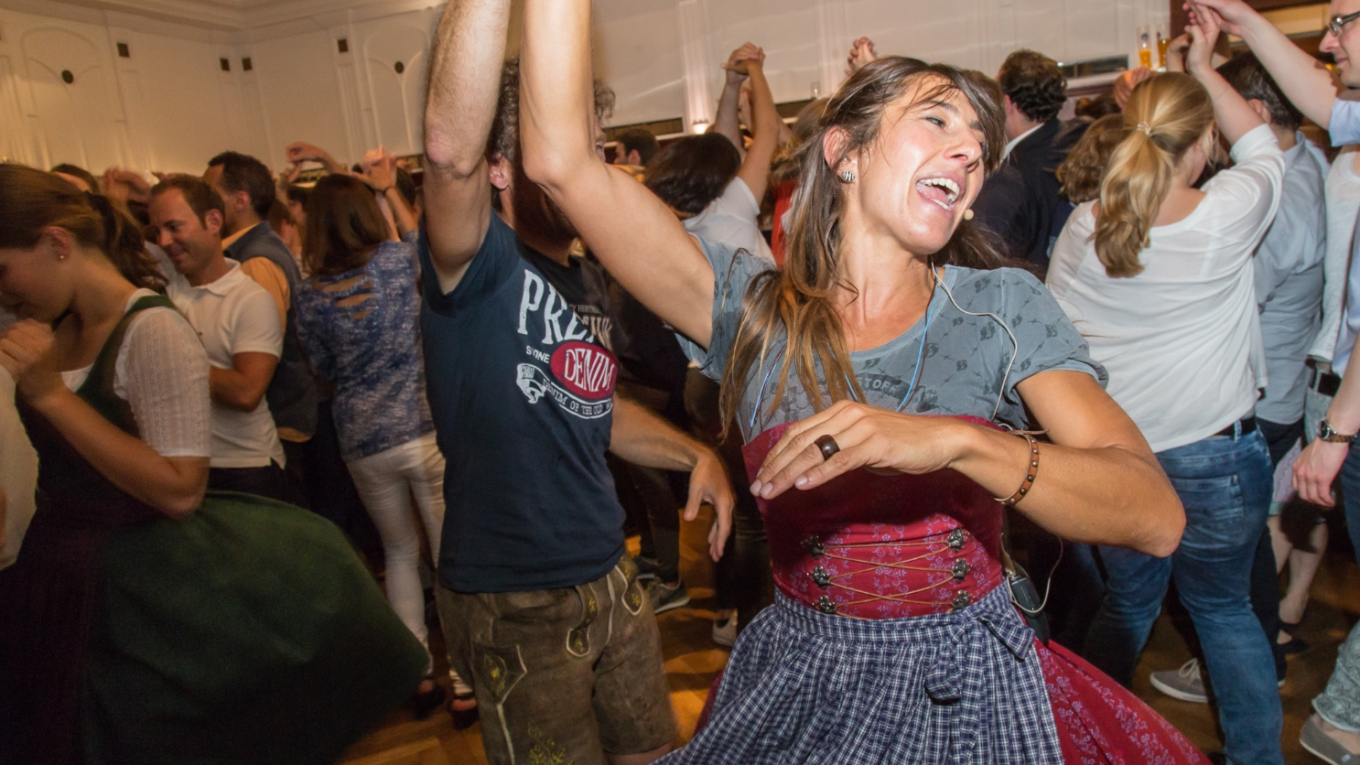 Im Vordergrund die Tanzmeisterin Katharina Mayer im rosa Dirndl. Sie bewegt sich mit gehobenen Armen zur Musik. Hinter ihr viele weitere Menschen, die ausgelassen tanzen.