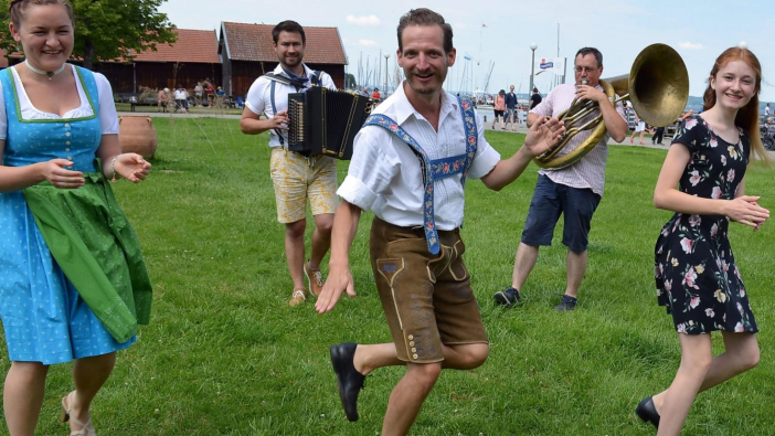 Auf einer Wiese ganzen junge Menschen in Tracht mit Blick in die Kamera, im Hintergrund spielt ein Mann Ziach, ein anderer Helikon.