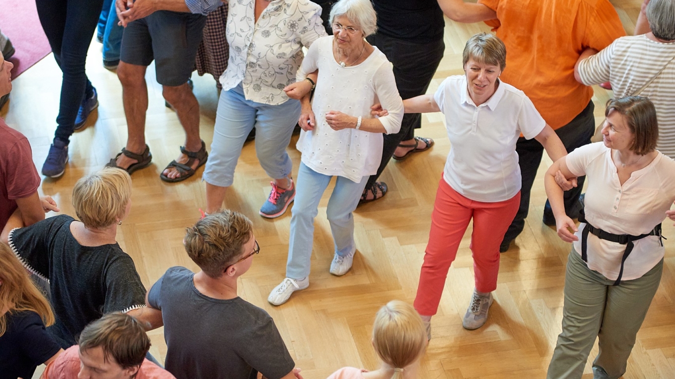 Menschen in Freizeitkleidung tanzen in einem Saal in Reihen eingehakt aufeinander zu.