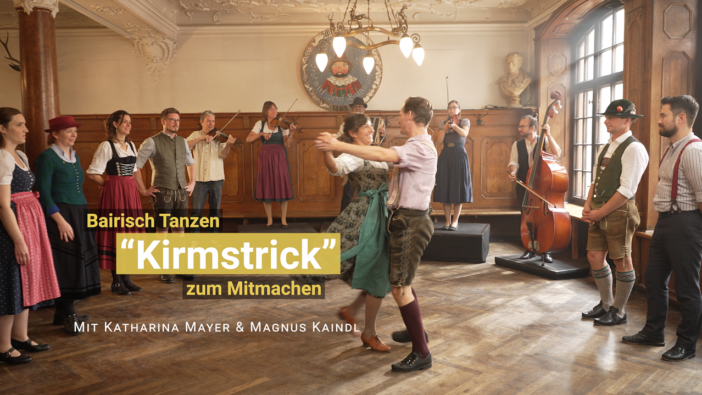 Ein Tanzpaar in Tracht tanzen einen Tanz vor. Andere Tänzerinnen und Tänzer schauen zu. Im Hintergrund spielt Livemusik.