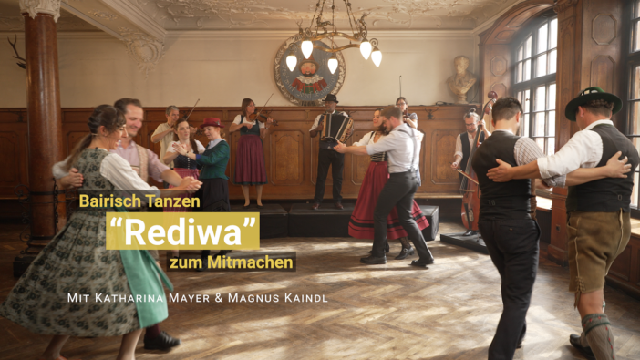 Vier Tanzpaare im bayerischer Kleidung drehen sich miteinander im Kreis. Im Hintergrund spielt Livemusik.