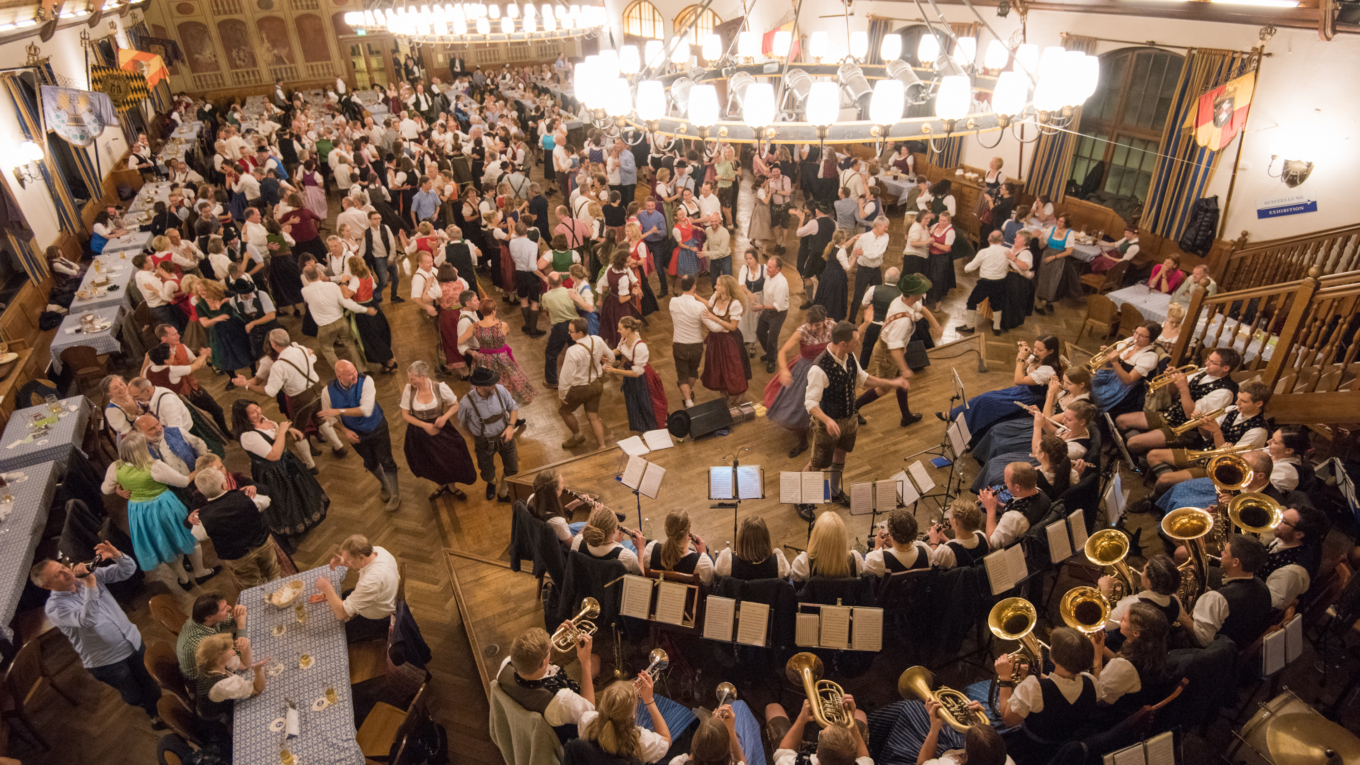 Blick in einen großen Saal mit vielen Tanzpaaren in Dirndl und Lederhosen und einer Blaskapelle auf der Bühne.